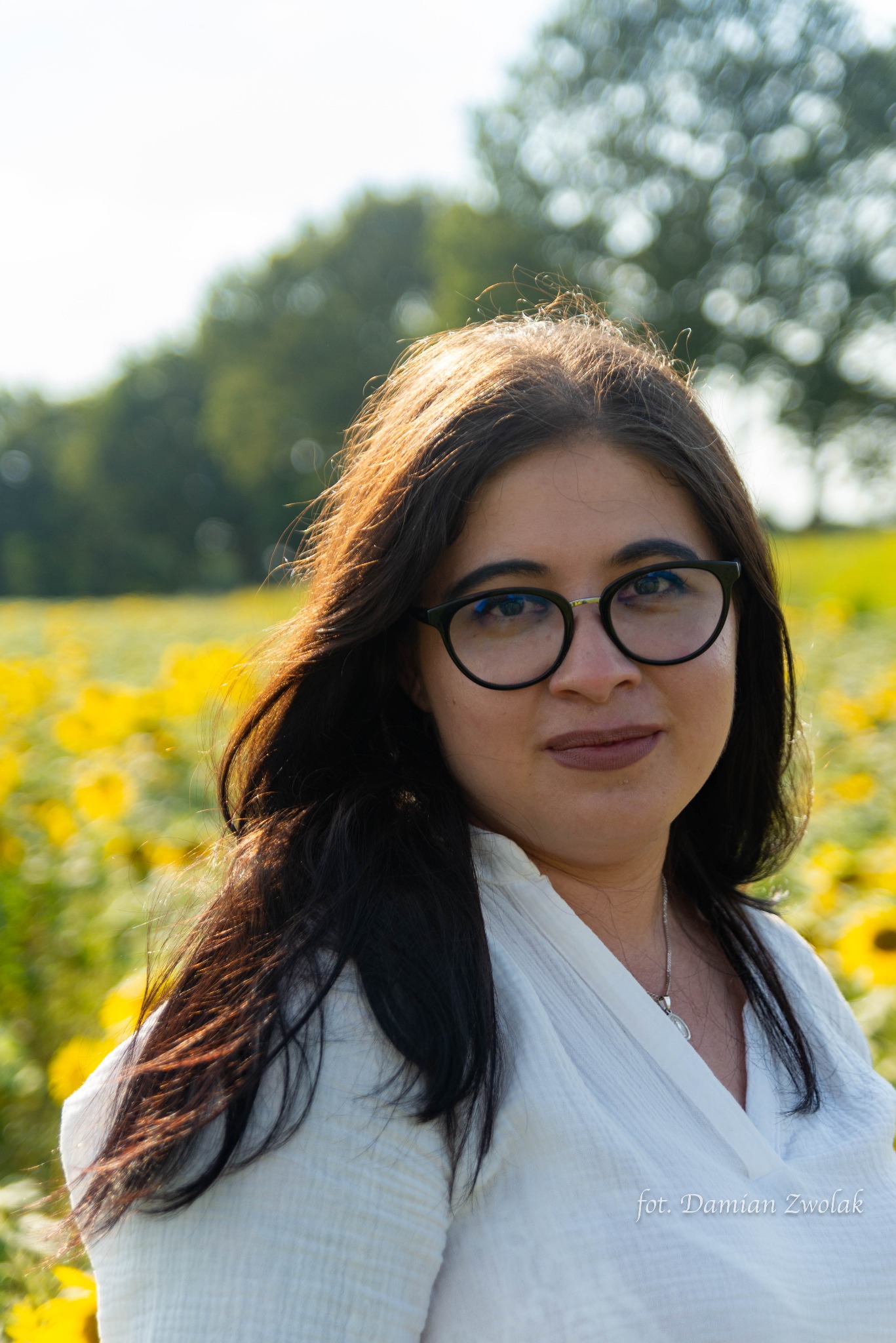 Portret kobiety w okularach na tle pola słoneczników w słoneczny dzień, ubrana w białą koszulę.