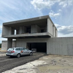 ALUSTAL SYSTEM Zamość Drzwi Okna Bramy Ogrodzenia - Surowy, dwukondygnacyjny dom z betonu komórkowego w trakcie budowy, z samochodem marki Volkswagen Passat z logo firmy ALUSTAL zaparkowanym na podjeździe z gruzem.