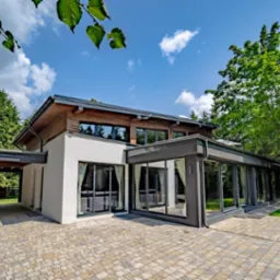 Nowoczesny dom jednorodzinny z dużymi oknami i przeszkloną werandą, widok z zewnątrz na starannie ułożoną kostkę brukową i zieleń ogrodu.