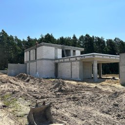 ALUSTAL SYSTEM Zamość Drzwi Okna Bramy Ogrodzenia - Budowa domu jednorodzinnego w stanie surowym otwartym, widoczne ściany z bloczków, betonowe nadproża, drabina oparta o ścianę, wykop pod fundamenty i koparka w tle.