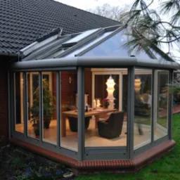 Szklana oranżeria z szarymi ramami okiennymi, przylegająca do ceglanego domu z ciemnym dachem, widok na stół i krzesła wewnątrz, oświetlone lampą i świecami.