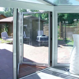 Otwarta, składana szklana ściana w oranżerii, widok na taras z meblami ogrodowymi i drewnianą altaną w tle.