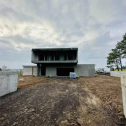 Surowy, dwukondygnacyjny dom w trakcie budowy z widocznymi oknami i otwartym wjazdem do garażu; na pierwszym planie teren budowy z workami materiałów budowlanych, w tle zaparkowane samochody...