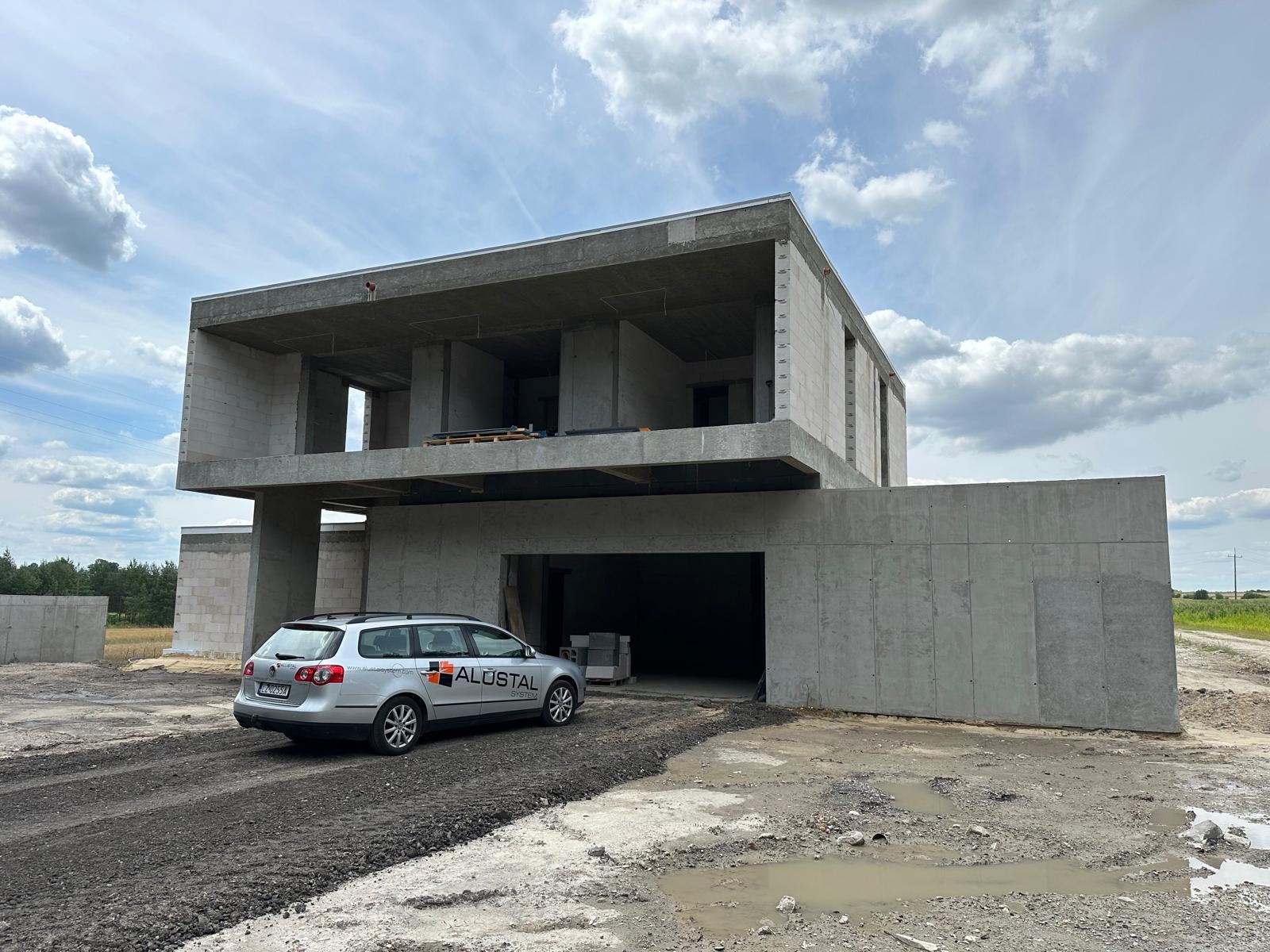Surowy, dwukondygnacyjny dom z betonu komórkowego w trakcie budowy, z samochodem marki Volkswagen Passat z logo firmy ALUSTAL zaparkowanym na podjeździe z gruzem.