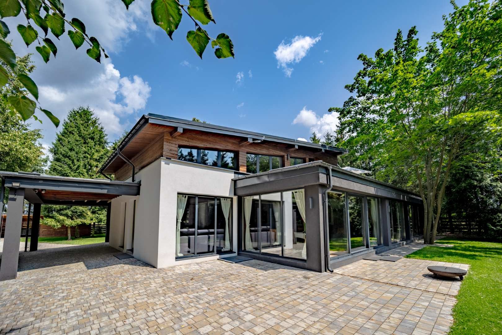 Nowoczesny dom jednorodzinny z dużymi oknami i przeszkloną werandą, widok z zewnątrz na starannie ułożoną kostkę brukową i zieleń ogrodu.