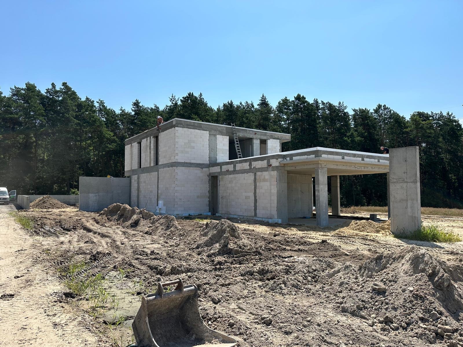 Budowa domu jednorodzinnego w stanie surowym otwartym, widoczne ściany z bloczków, betonowe nadproża, drabina oparta o ścianę, wykop pod fundamenty i koparka w tle.