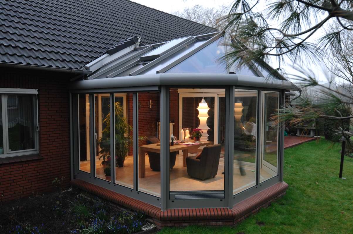Szklana oranżeria z szarymi ramami okiennymi, przylegająca do ceglanego domu z ciemnym dachem, widok na stół i krzesła wewnątrz, oświetlone lampą i świecami.