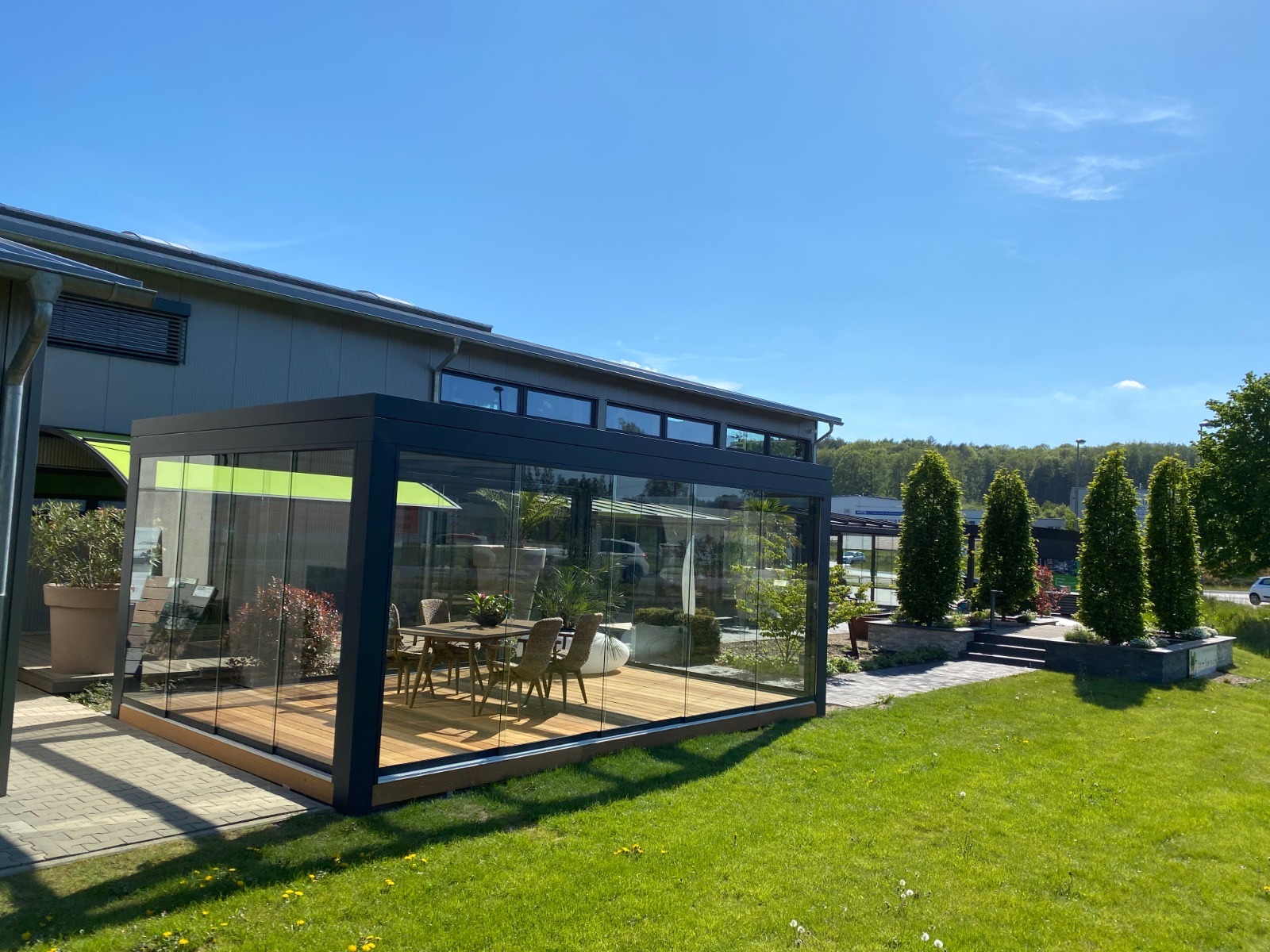 Szklana pergola z drewnianą podłogą i meblami ogrodowymi, widok z zewnątrz na tle zielonego trawnika i błękitnego nieba.