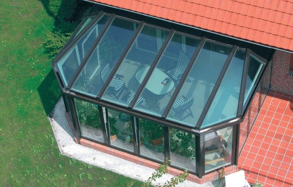 Zadaszony taras z przeszklonymi ścianami i dachem, widok z góry, na podłodze czerwona terakota, wewnątrz stolik i krzesła w paski.