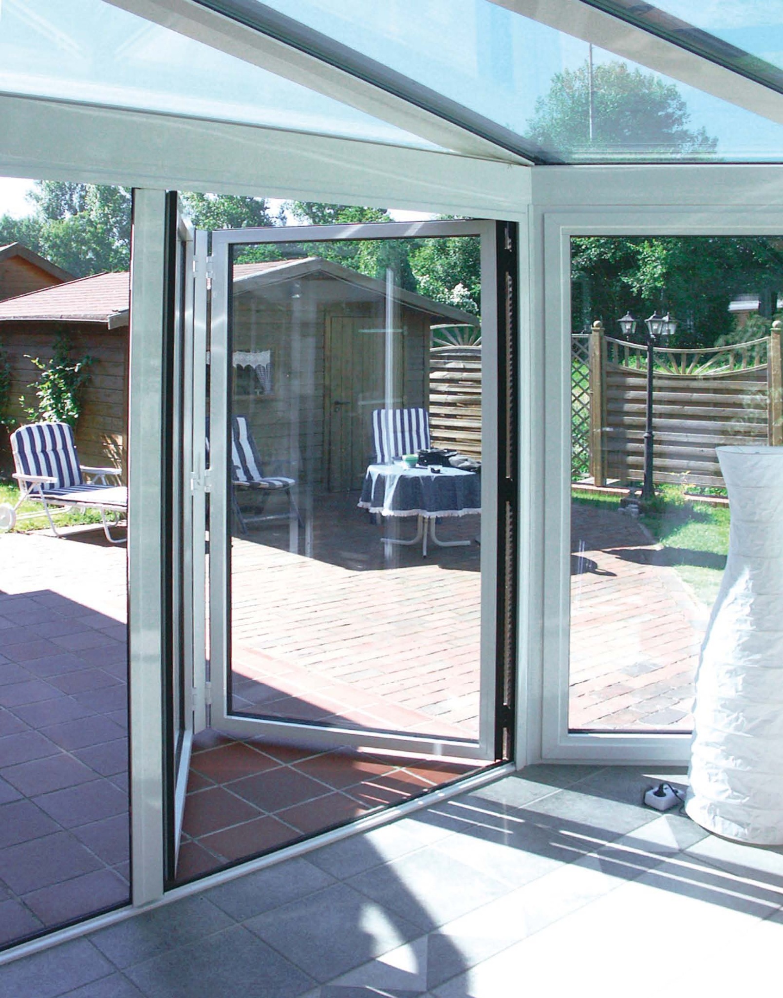 Otwarta, składana szklana ściana w oranżerii, widok na taras z meblami ogrodowymi i drewnianą altaną w tle.