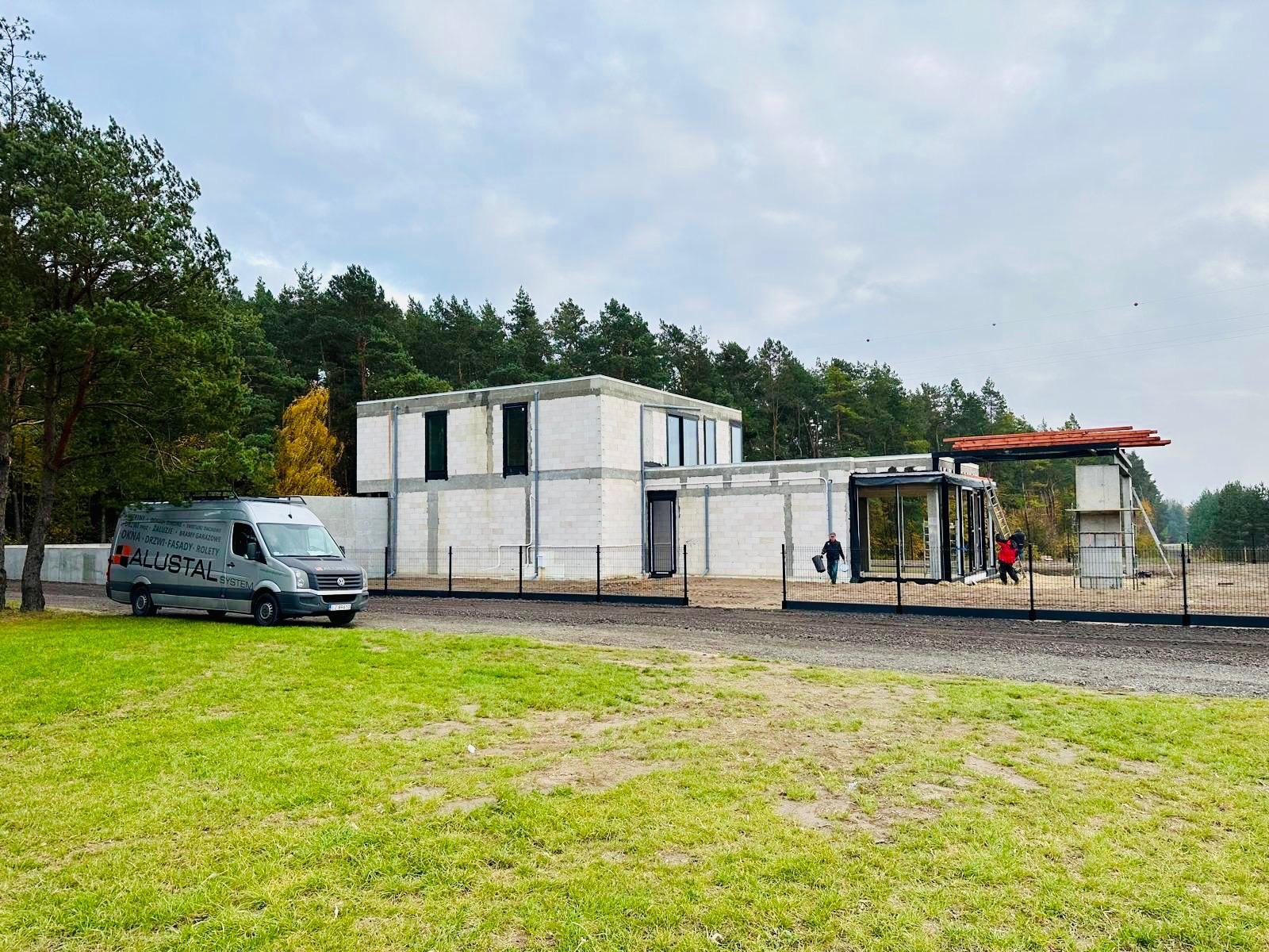 Budowa nowoczesnego domu jednorodzinnego w Zamościu z zamontowanymi oknami i drzwiami, widoczny samochód dostawczy firmy Alustal oraz pracownicy na budowie.