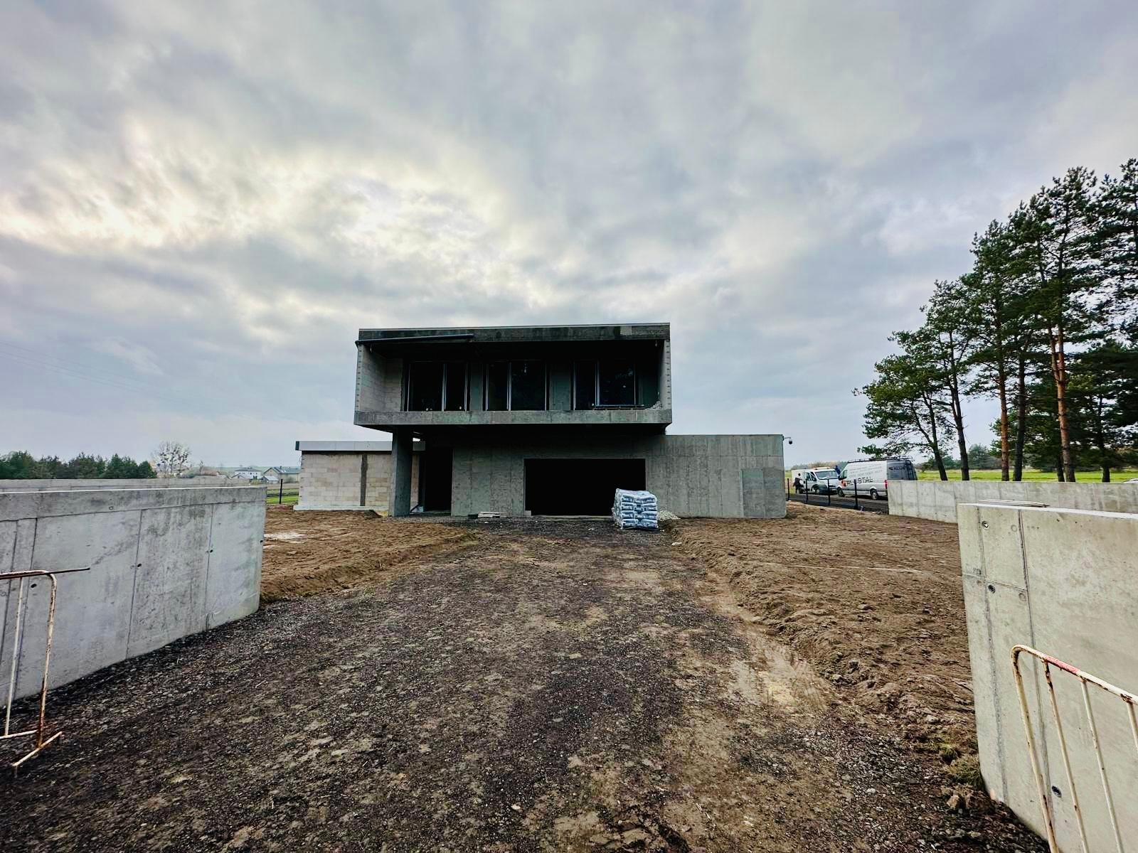 Surowy, dwukondygnacyjny dom w trakcie budowy z widocznymi oknami i otwartym wjazdem do garażu; na pierwszym planie teren budowy z workami materiałów budowlanych, w tle zaparkowane samochody...