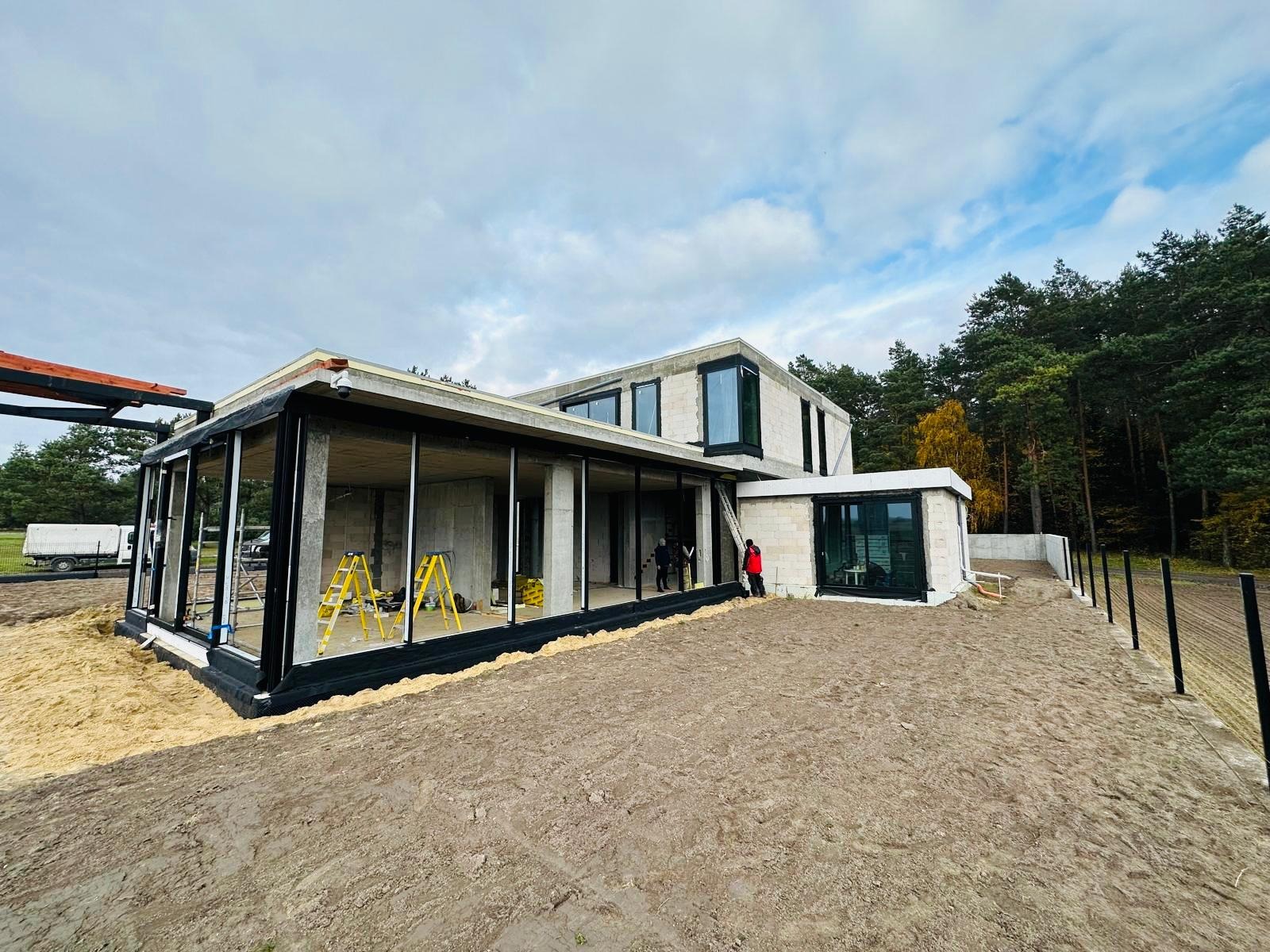 Surowy, dwukondygnacyjny dom w trakcie budowy z dużymi przeszkleniami, widoczne żółte drabiny i pracownicy, teren wokół domu w stanie przygotowania pod trawnik, las w tle.