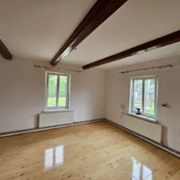 Puste wnętrze pokoju z drewnianą podłogą, białymi ścianami, dwoma oknami z widokiem na zieleń i odsłoniętymi, ciemnymi belkami stropowymi.