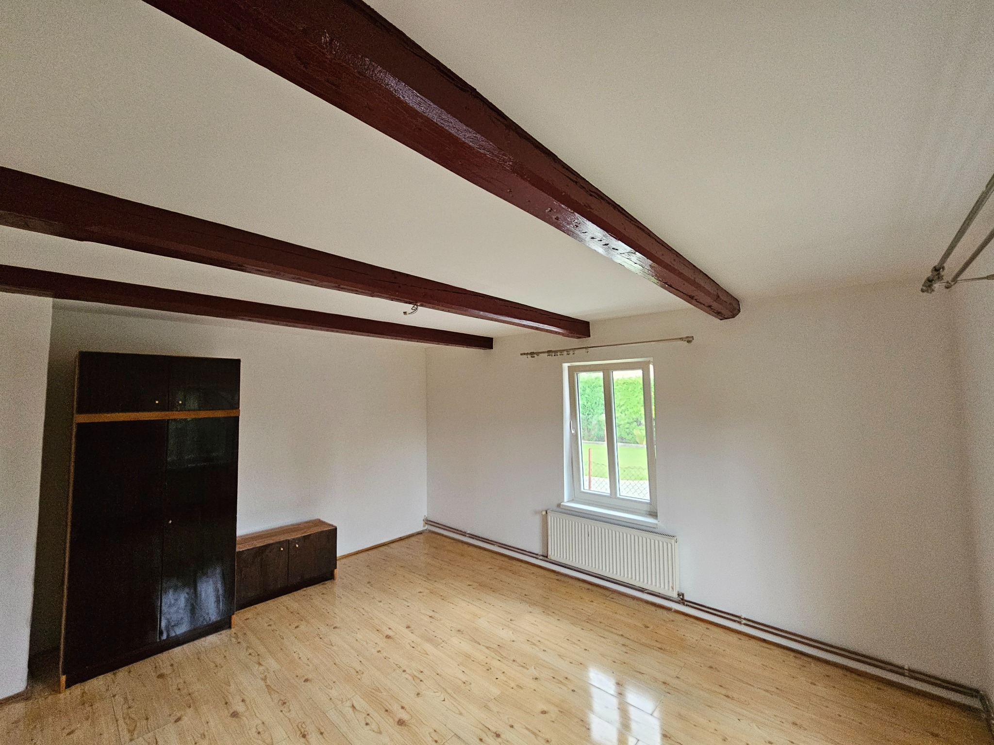 Puste, jasne pomieszczenie z drewnianą podłogą, ciemnymi belkami stropowymi, grzejnikiem pod oknem i ciemnymi meblami przy ścianie.