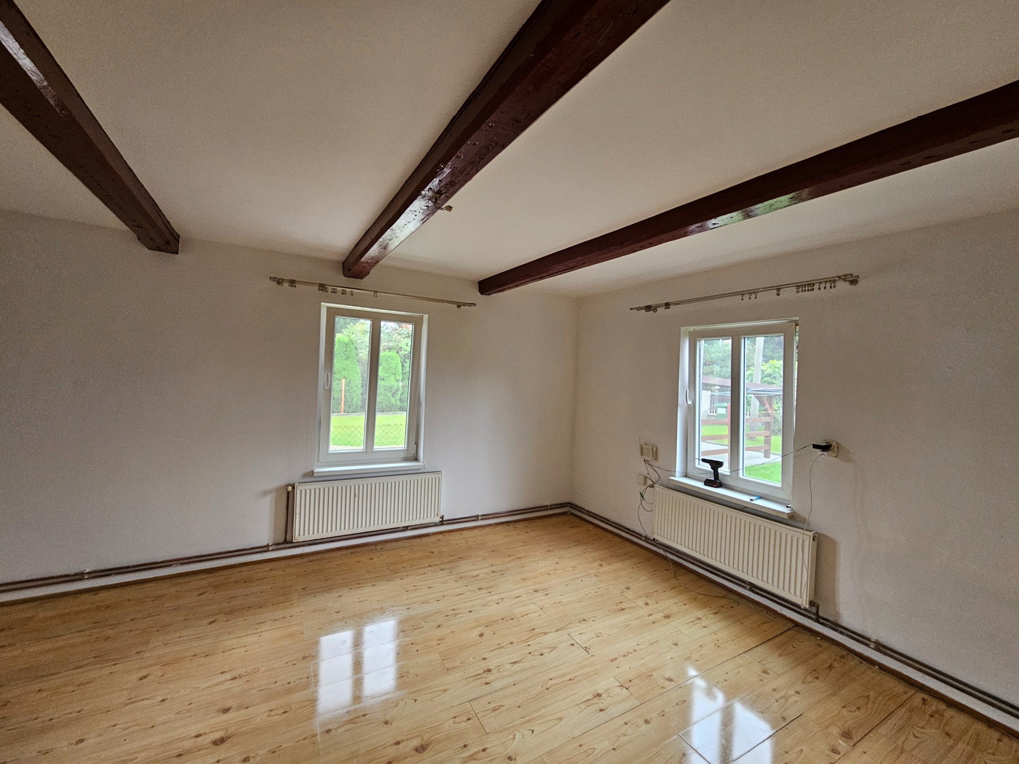 Puste wnętrze pokoju z drewnianą podłogą, białymi ścianami, dwoma oknami z widokiem na zieleń i odsłoniętymi, ciemnymi belkami stropowymi.