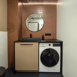 BK Design Barbara Kubica - Nowoczesna łazienka z okrągłym lustrem, umywalką nablatową i pralką. Ściana w odcieniach brązu z subtelnym oświetleniem LED. Minimalistyczny design i funkcjonalne rozwiązania.