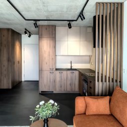 BK Design Barbara Kubica - Nowoczesna kuchnia z drewnianymi szafkami, czarnym blatem i oświetleniem punktowym. W tle widoczny salon z pomarańczową sofą i stolikiem kawowym z kwiatami.