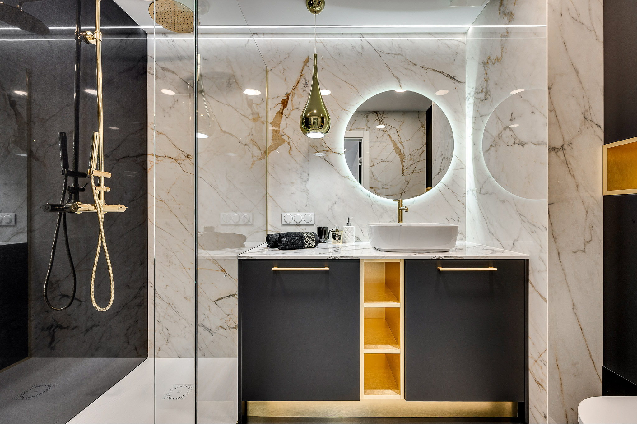 Elegancka łazienka z marmurowymi płytkami, okrągłym lustrem z podświetleniem LED i złotymi akcentami. Kabina prysznicowa ze złotą armaturą.