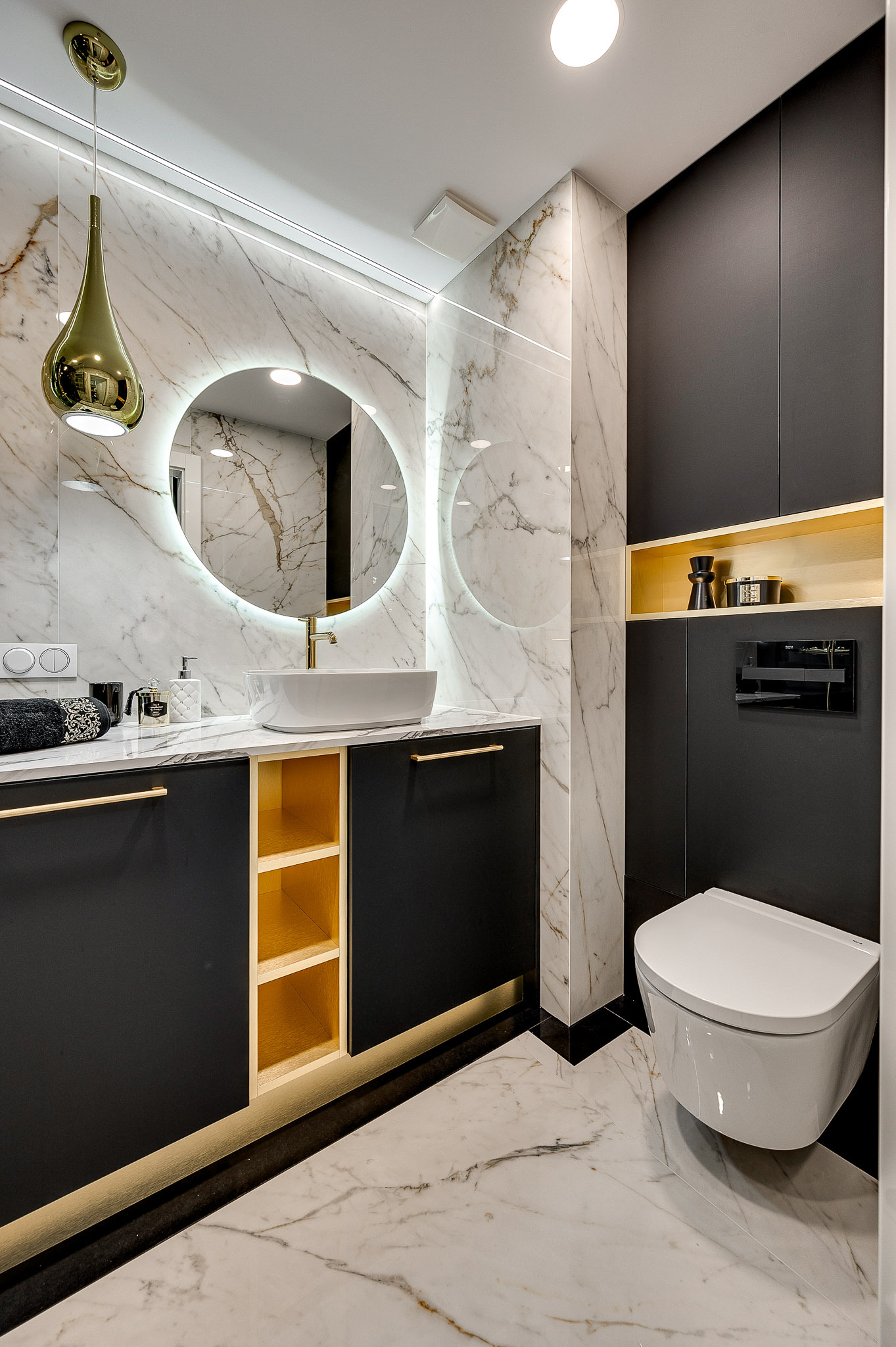 Elegancka łazienka z marmurowymi płytkami, okrągłym lustrem z podświetleniem LED, czarnymi szafkami z złotymi akcentami i wiszącą toaletą. Nowoczesny design.