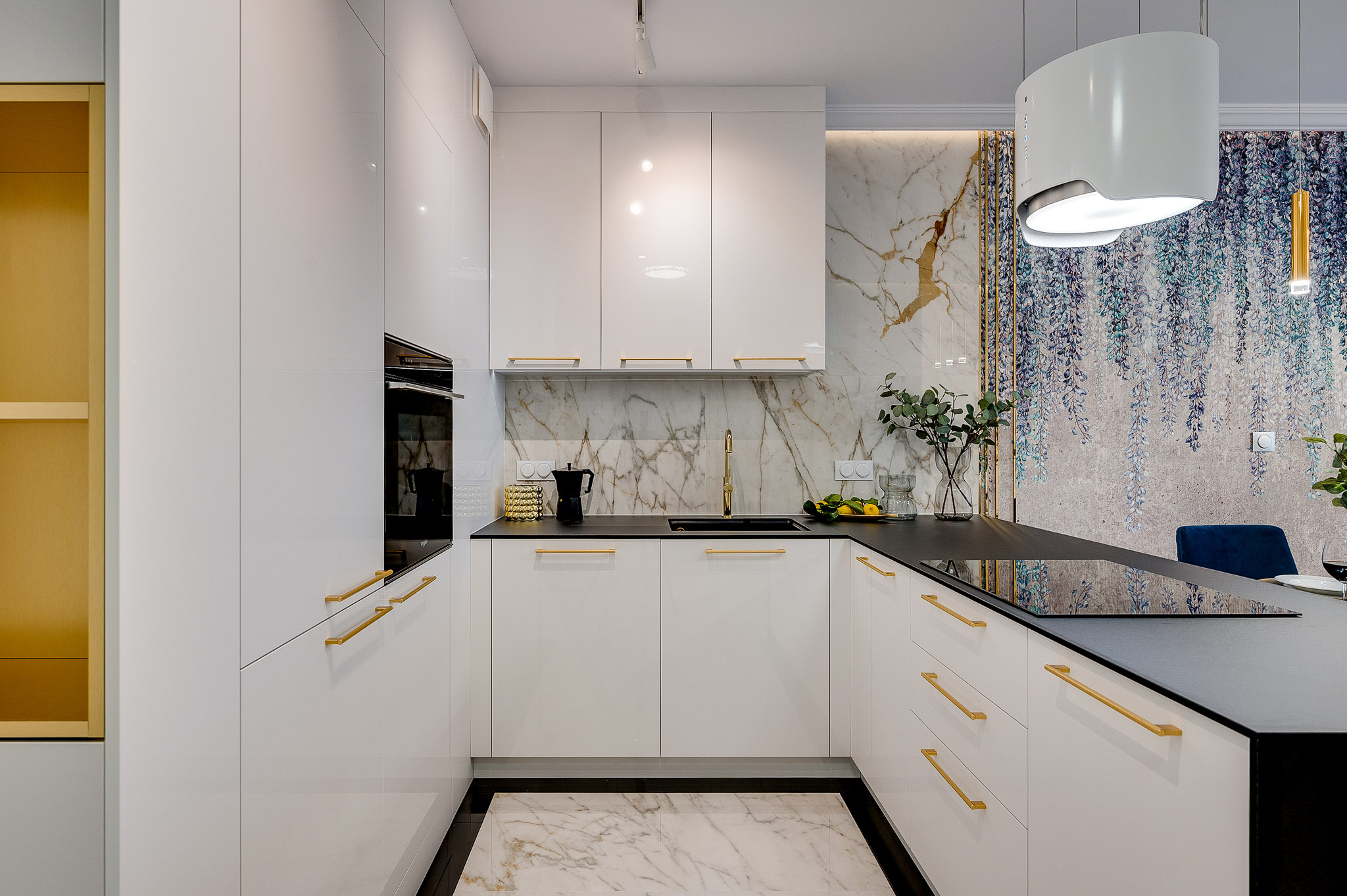 Nowoczesna kuchnia w bieli z złotymi akcentami: marmurowy backsplash, czarny blat, designerski okap i dekoracyjna tapeta z motywem roślinnym. Eleganckie wykończenie.