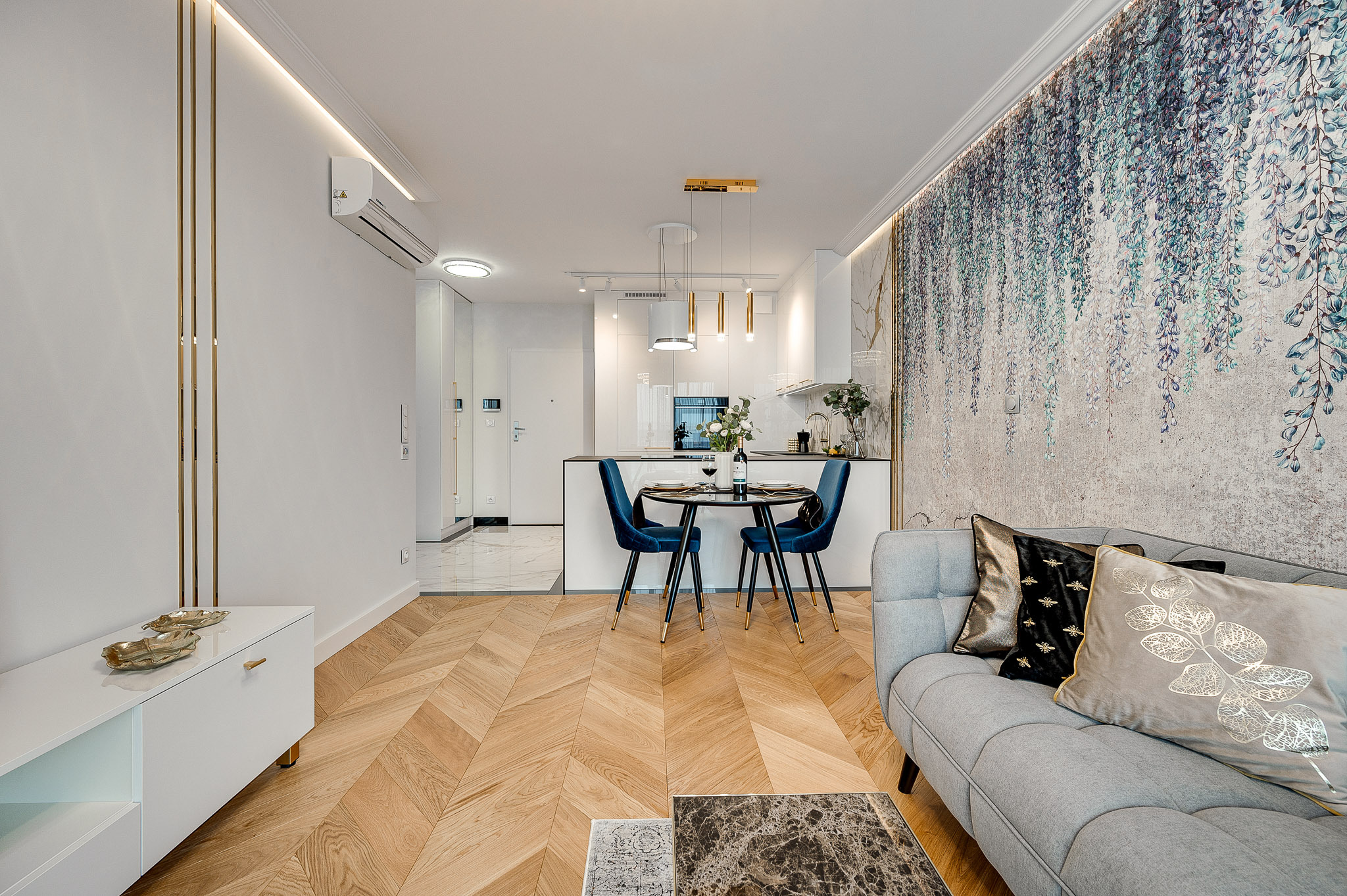 Nowoczesne wnętrze: parkiet w jodełkę, sofa z poduszkami, okrągły stół z krzesłami i dekoracyjna tapeta z motywem roślinnym. Minimalistyczna kuchnia w tle.