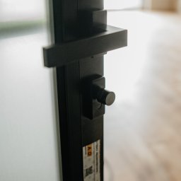 FINESTRA S.C. DANIEL LULEWICZ, MICHAŁ KULESZA - Detal czarnej klamki i zamka w minimalistycznych drzwiach z białym wypełnieniem, widoczne naklejki producenta na ramie, w tle jasne pomieszczenie.