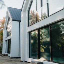 FINESTRA S.C. DANIEL LULEWICZ, MICHAŁ KULESZA - Nowoczesny dom z dużymi oknami aluminiowymi, odbijającymi drzewa. Biała elewacja, szary dach. Widok z zewnątrz, teren w trakcie zagospodarowania.