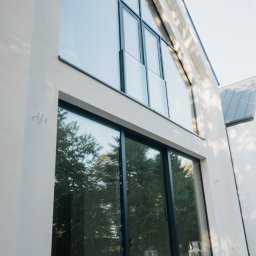 FINESTRA S.C. DANIEL LULEWICZ, MICHAŁ KULESZA - Nowoczesna fasada domu z dużymi oknami aluminiowymi w czarnych ramach, odbijającymi zieleń drzew. Na dole paleta, sugerująca trwające prace wykończeniowe.
