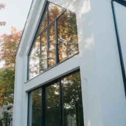 Nowoczesny dom z dużymi oknami aluminiowymi, w których odbija się jesienne drzewo. Widok z dołu, podkreślający geometrię budynku i okien.