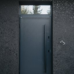 FINESTRA S.C. DANIEL LULEWICZ, MICHAŁ KULESZA - Ciemnoszare drzwi wejściowe z długim uchwytem i górnym oknem, wkomponowane w ciemną, teksturowaną elewację budynku. Minimalistyczny design.