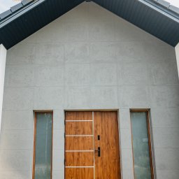 FINESTRA S.C. DANIEL LULEWICZ, MICHAŁ KULESZA - Nowoczesny dom z drewnianymi drzwiami wejściowymi i wąskimi oknami po bokach. Elewacja w odcieniach szarości, dach dwuspadowy z ciemnej dachówki.