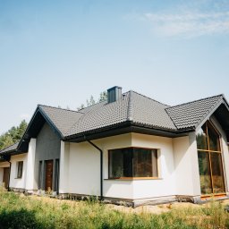 FINESTRA S.C. DANIEL LULEWICZ, MICHAŁ KULESZA - Nowoczesny dom jednorodzinny z dużymi oknami i szarą dachówką, otoczony zielenią. Widoczny fragment elewacji z oknami i drzwiami wejściowymi.