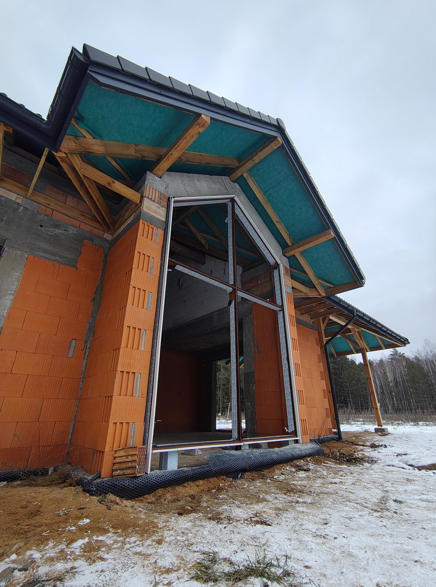 Budynek w trakcie budowy z widoczną ramą okienną, cegłami ceramicznymi i drewnianą konstrukcją dachu pod zieloną membraną.