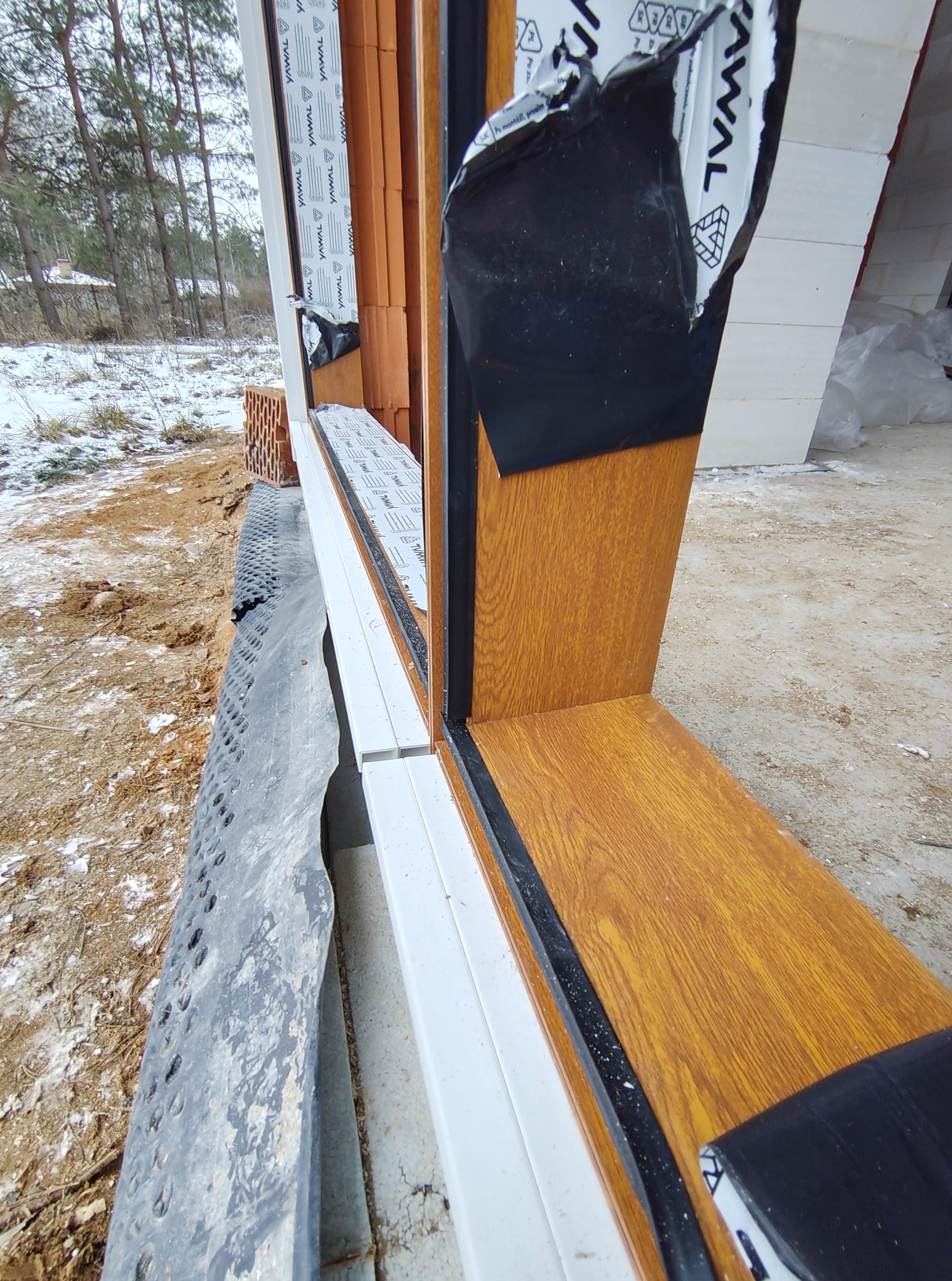 Fragment zamontowanego okna z drewnopodobną ramą, widok z zewnątrz na surową ścianę z cegieł i folię ochronną na profilu.