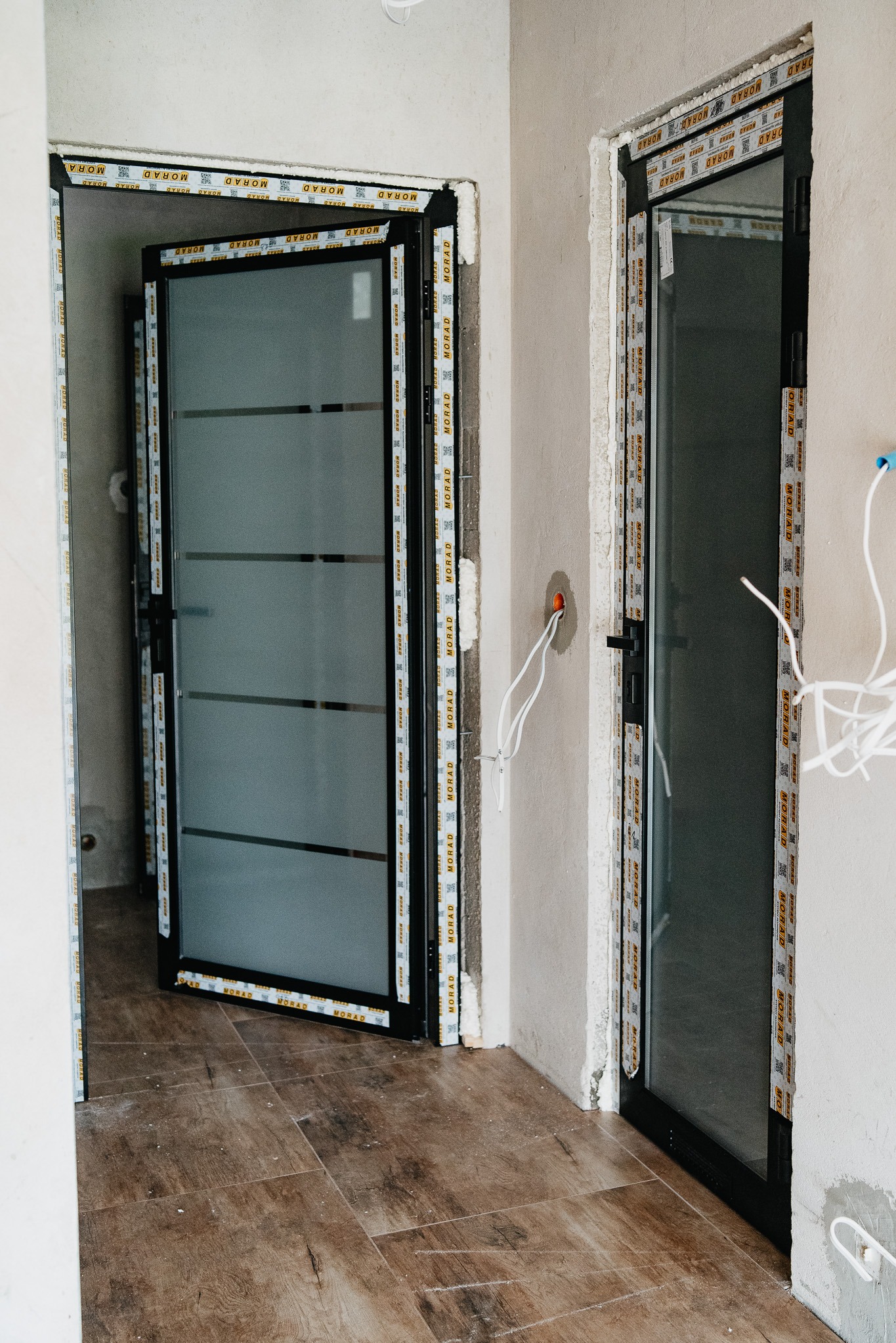 Montaż czarnych drzwi przesuwnych z matowego szkła z ramą aluminiową, widoczne pianowanie uszczelniające ościeżnicę, instalacja elektryczna w ścianie, podłoga wyłożona płytkami imitującymi drewno.