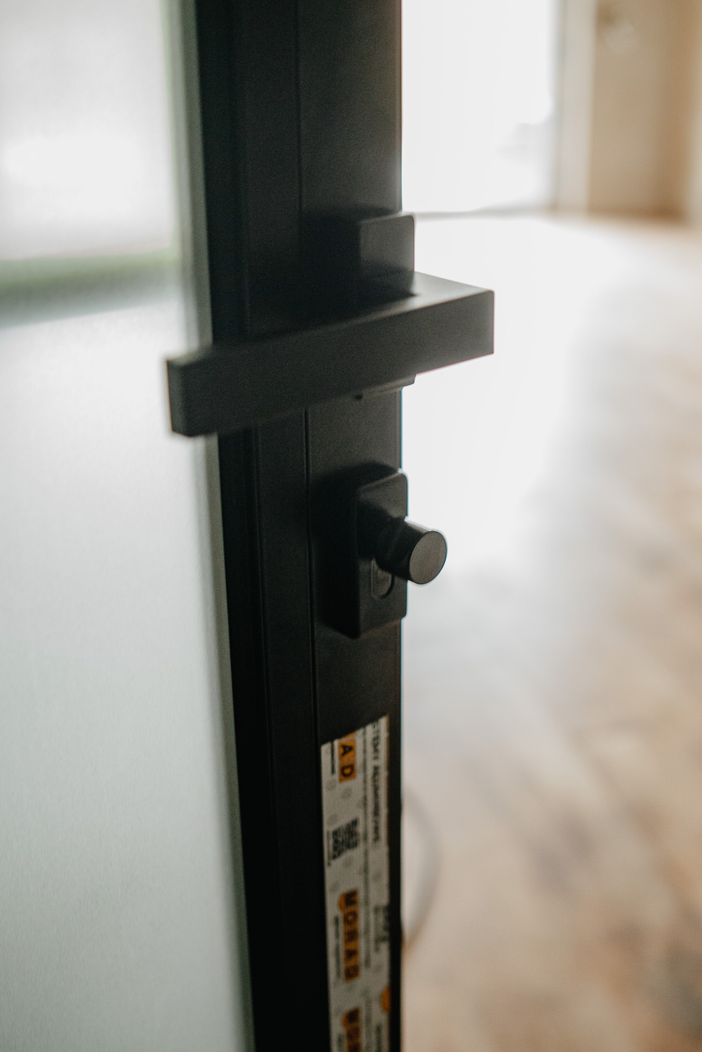 Detal czarnej klamki i zamka w minimalistycznych drzwiach z białym wypełnieniem, widoczne naklejki producenta na ramie, w tle jasne pomieszczenie.