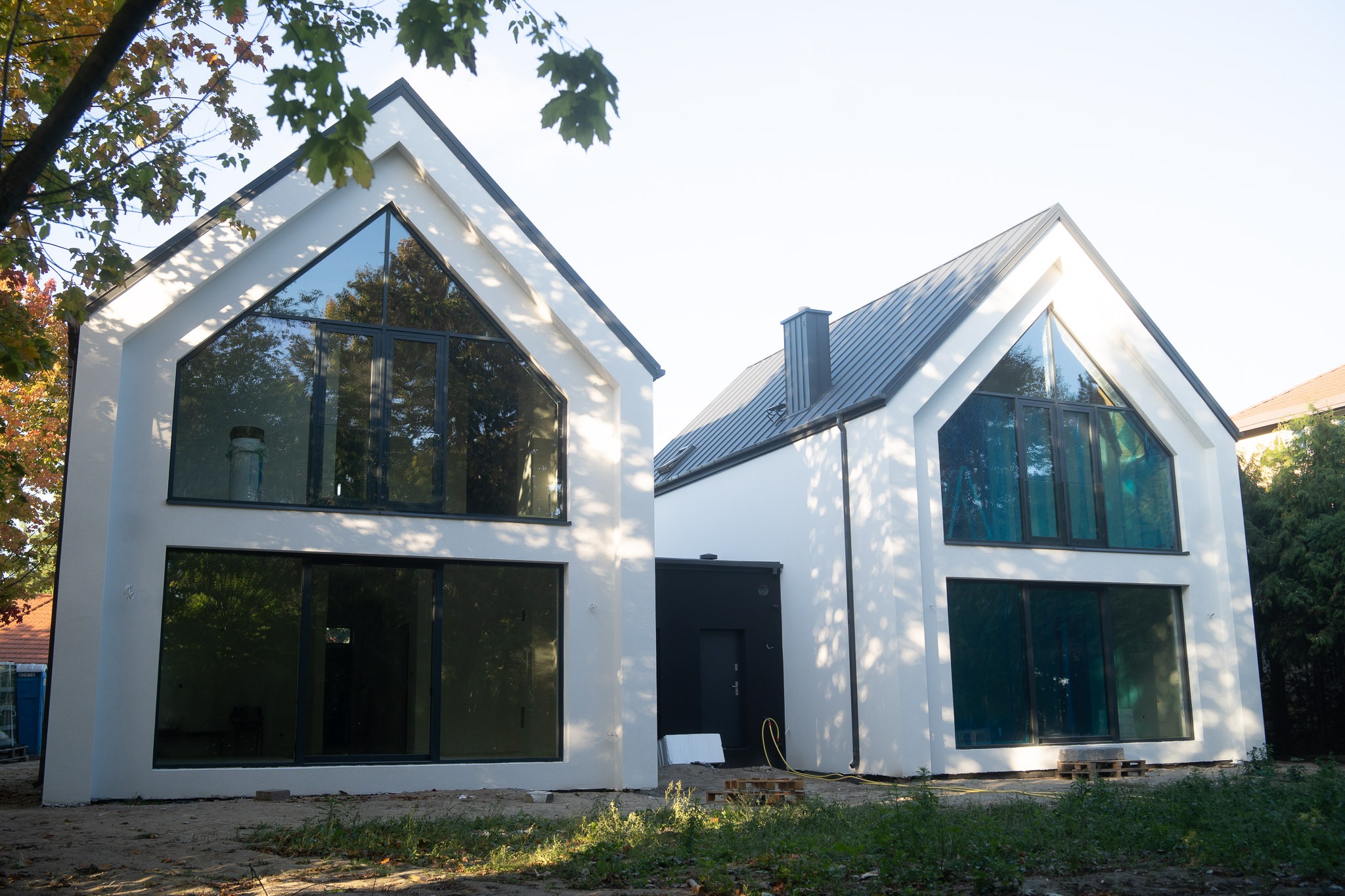 Dwa nowoczesne, białe domy z dużymi oknami i ciemnymi ramami, połączone czarnym łącznikiem. Jeden dom z dachem dwuspadowym, drugi z dachem skośnym. Jesienne drzewa w tle.
