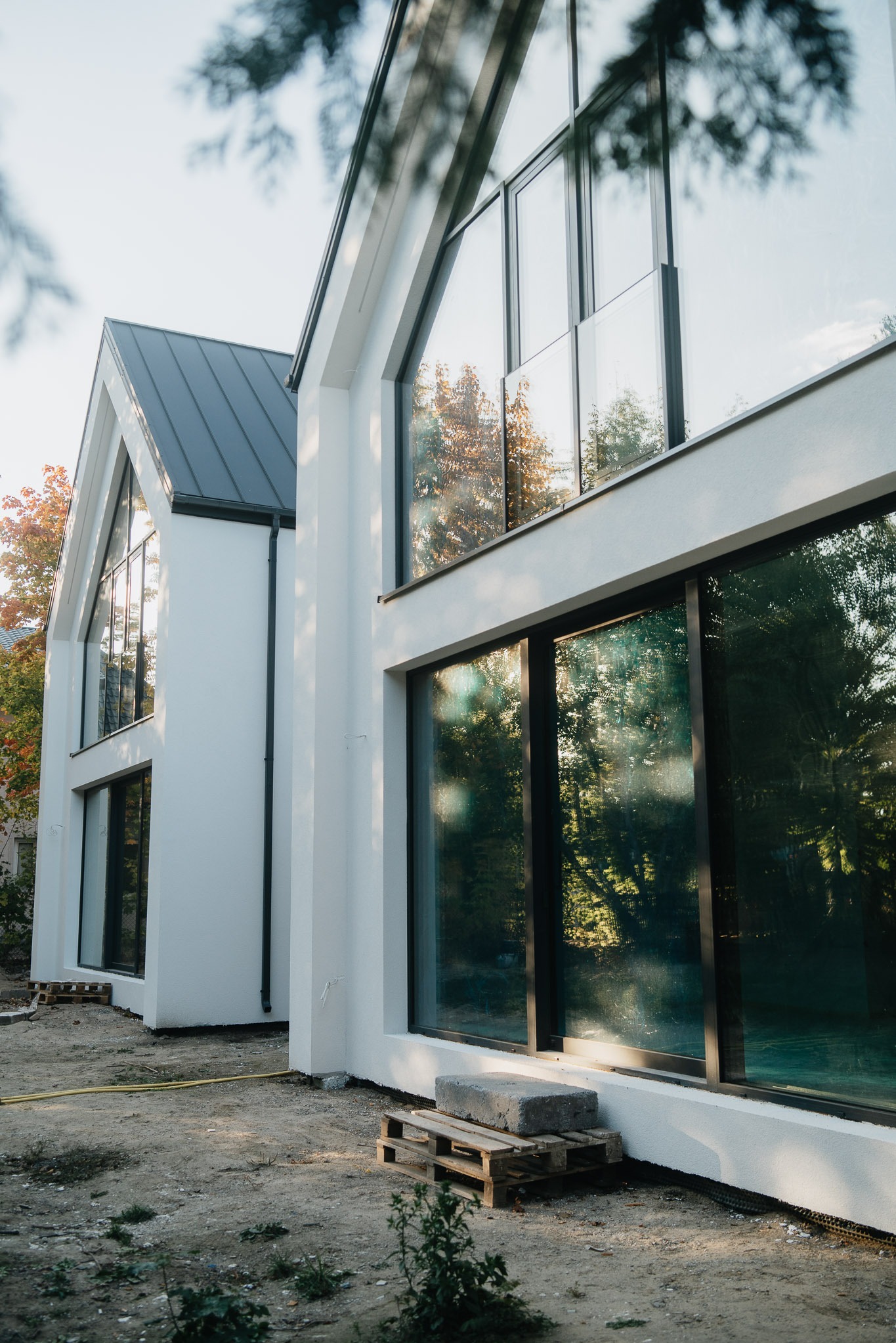 Nowoczesny dom z dużymi oknami aluminiowymi, odbijającymi drzewa. Biała elewacja, szary dach. Widok z zewnątrz, teren w trakcie zagospodarowania.
