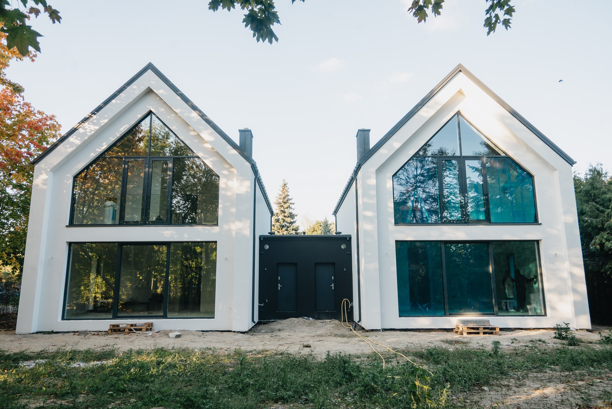 Dwa nowoczesne, białe domy z dużymi oknami, połączone czarnym łącznikiem z dwoma drzwiami. Widok z zewnątrz, w tle drzewa i błękitne niebo.