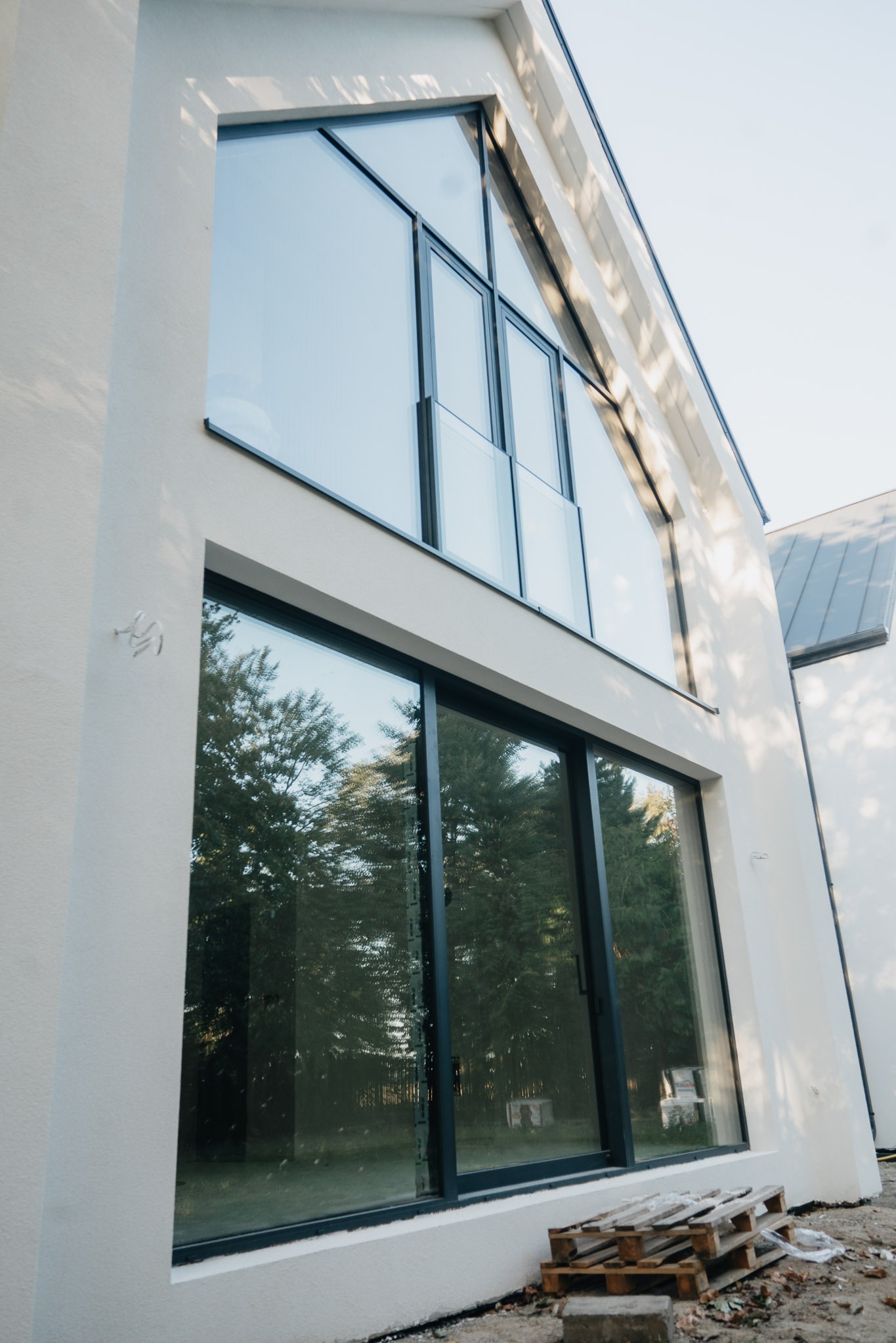 Nowoczesna fasada domu z dużymi oknami aluminiowymi w czarnych ramach, odbijającymi zieleń drzew. Na dole paleta, sugerująca trwające prace wykończeniowe.