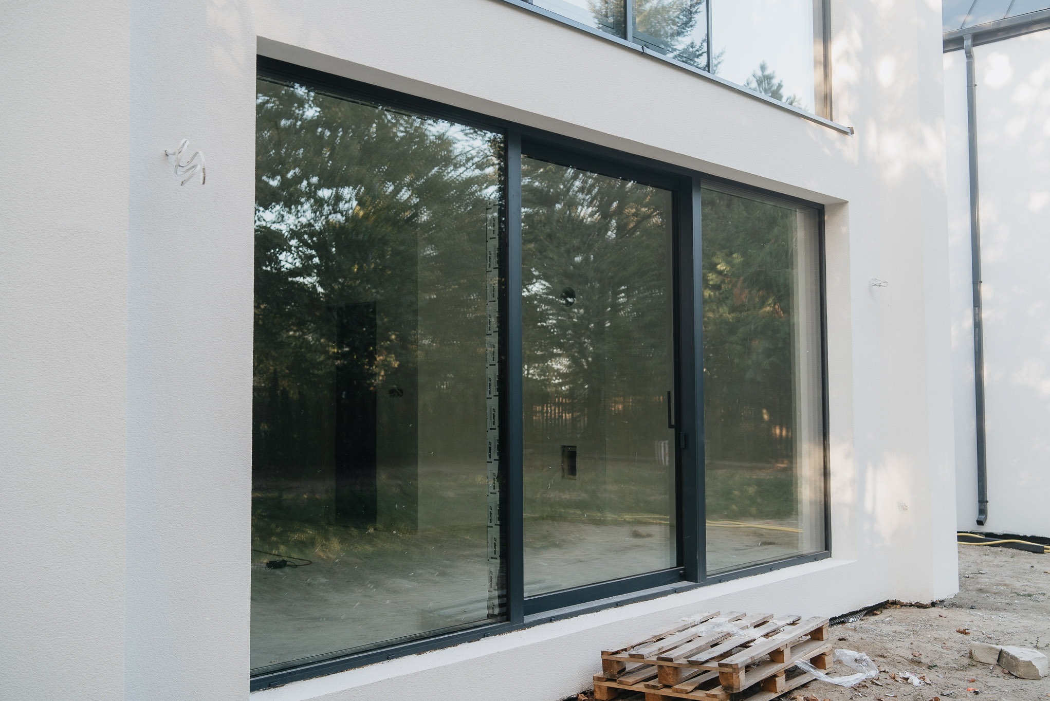 Duże, przesuwne okno aluminiowe w nowoczesnym domu z jasną elewacją. Widoczne drzewa odbijające się w szybie. Paleta leży na ziemi.