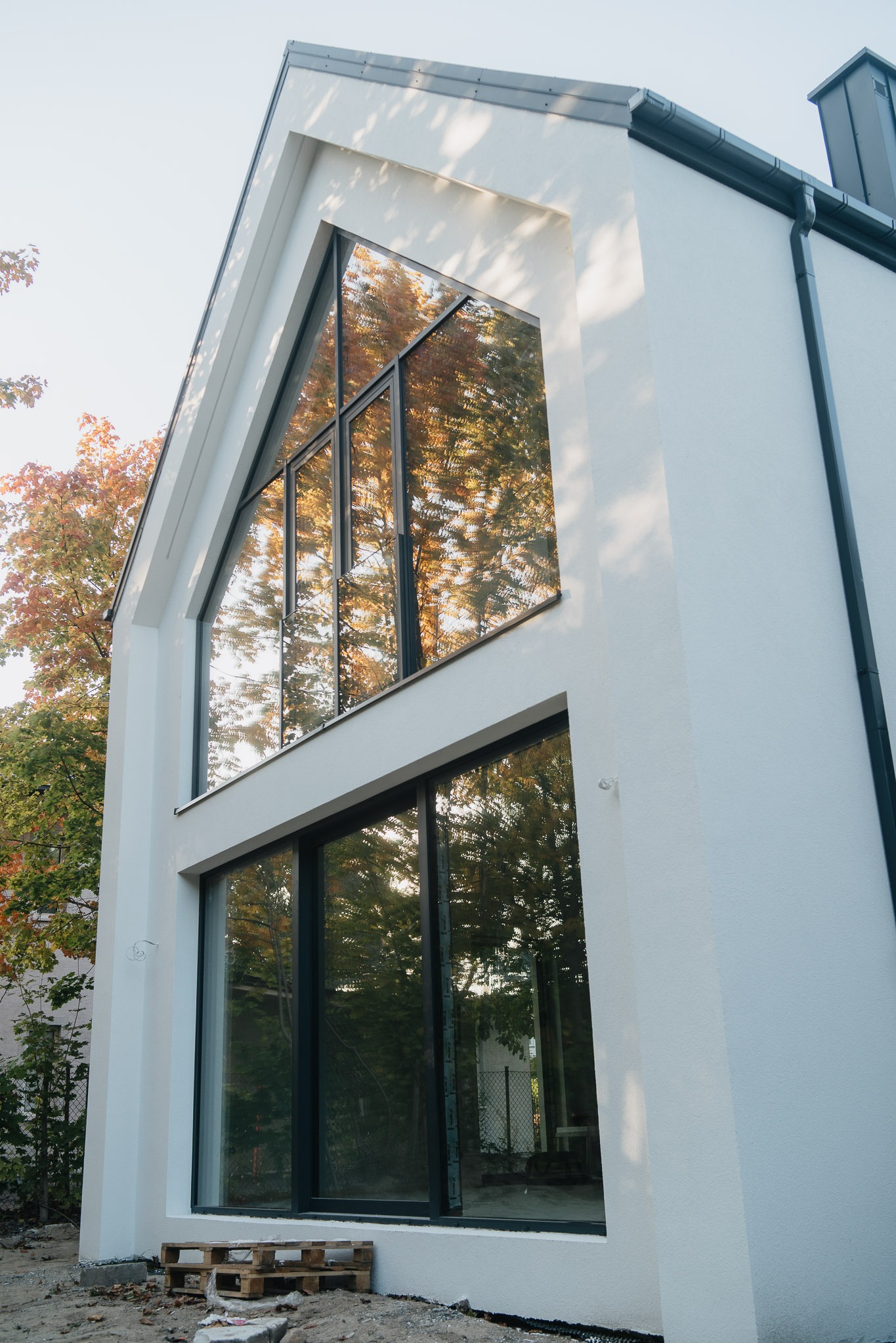 Nowoczesny dom z dużymi oknami aluminiowymi, w których odbija się jesienne drzewo. Widok z dołu, podkreślający geometrię budynku i okien.