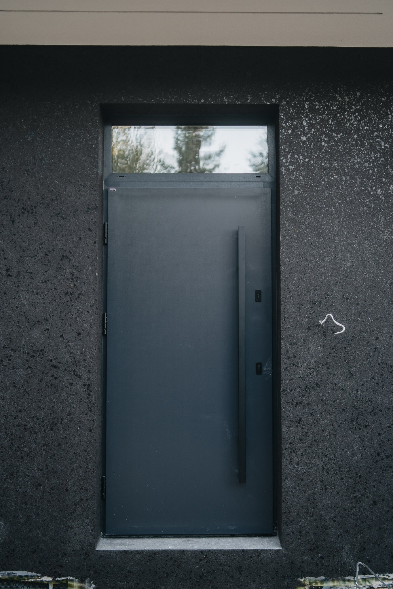 Ciemnoszare drzwi wejściowe z długim uchwytem i górnym oknem, wkomponowane w ciemną, teksturowaną elewację budynku. Minimalistyczny design.