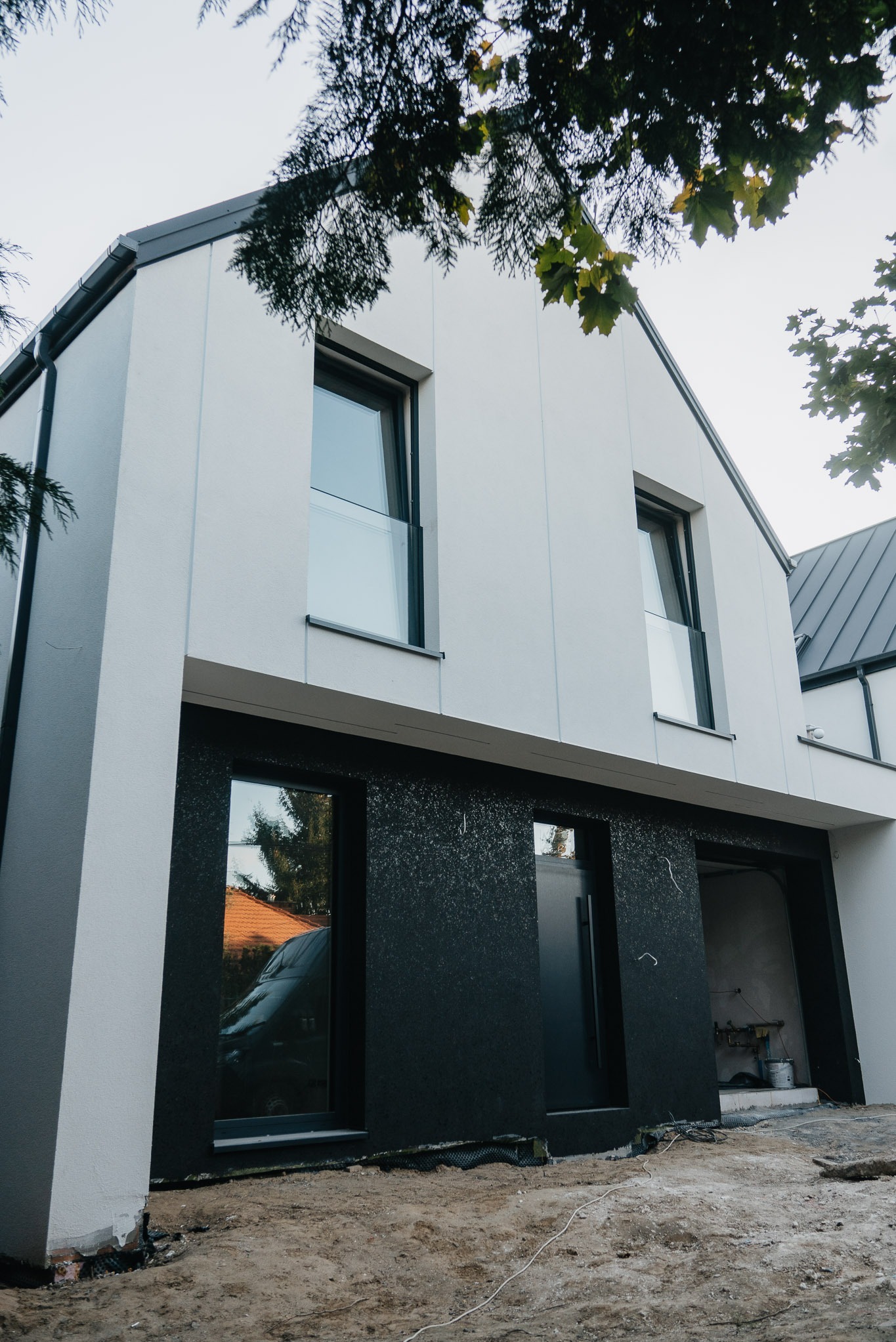 Nowoczesny dom z oknami i drzwiami w kolorze antracytu w Fasty. Widoczne okna na parterze z odbiciem otoczenia i drzwi wejściowe. Minimalistyczna architektura.