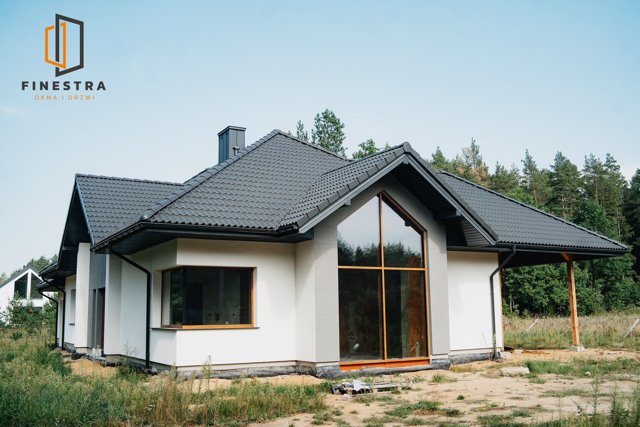 Nowoczesny dom jednorodzinny z dużym oknem szklanym w kształcie trójkąta, szara dachówka, jasna elewacja, widok z zewnątrz, trawa i drzewa w tle.