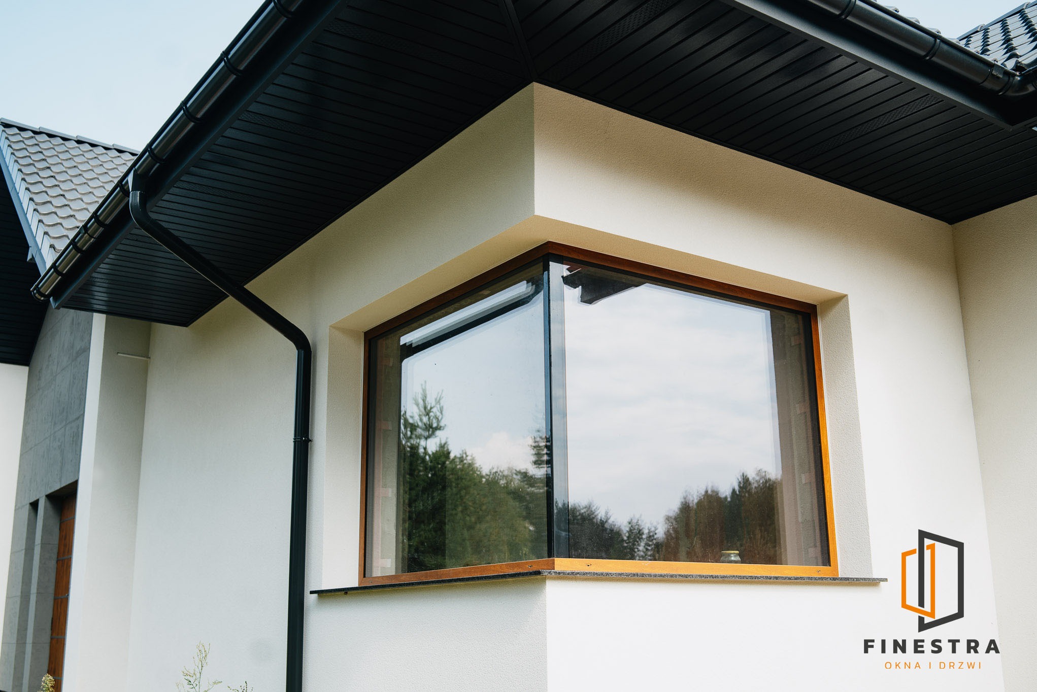 Narożne okno z drewnianą ramą w nowoczesnym domu z czarnym dachem i rynnami. Widok na zieleń za oknem. Logo firmy w prawym dolnym rogu.
