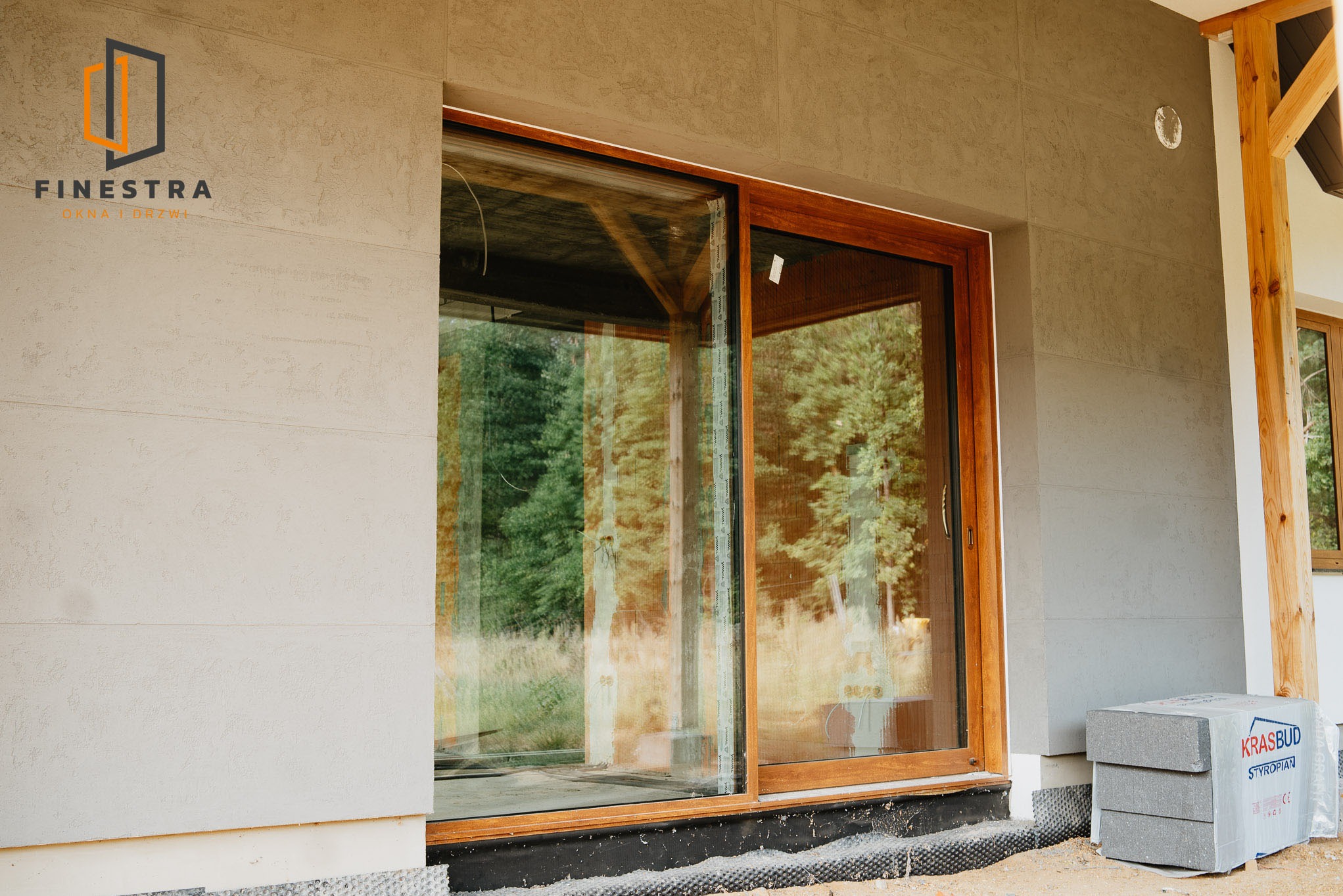 Duże, przesuwne okno z drewnianą ramą w elewacji budynku. Widoczne detale montażu i otoczenie domu. Na pierwszym planie paczka styropianu.