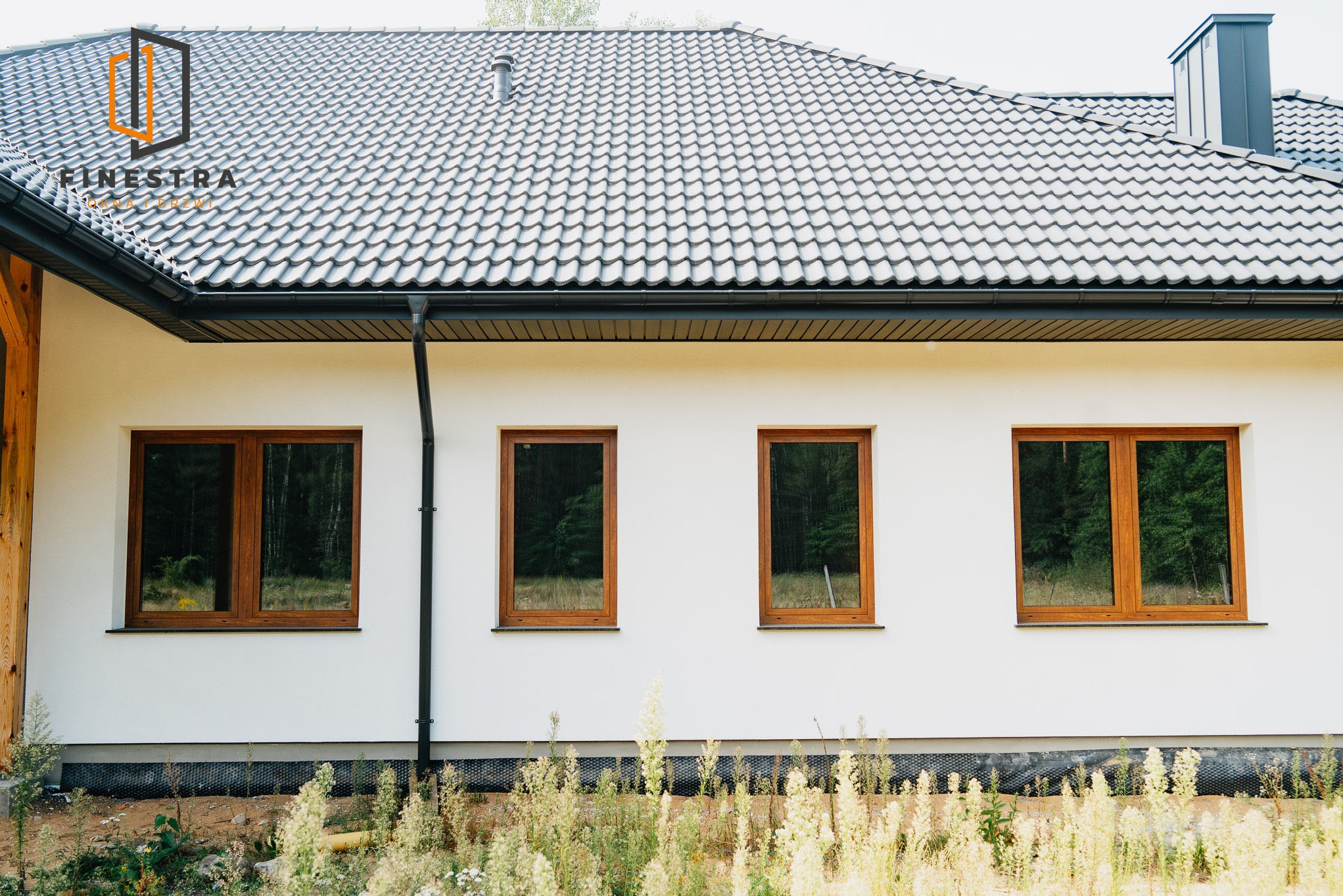 Elewacja domu z czterema drewnianymi oknami. Widoczne szare dachówki i biała ściana. W tle drzewa odbijające się w szybach okien.