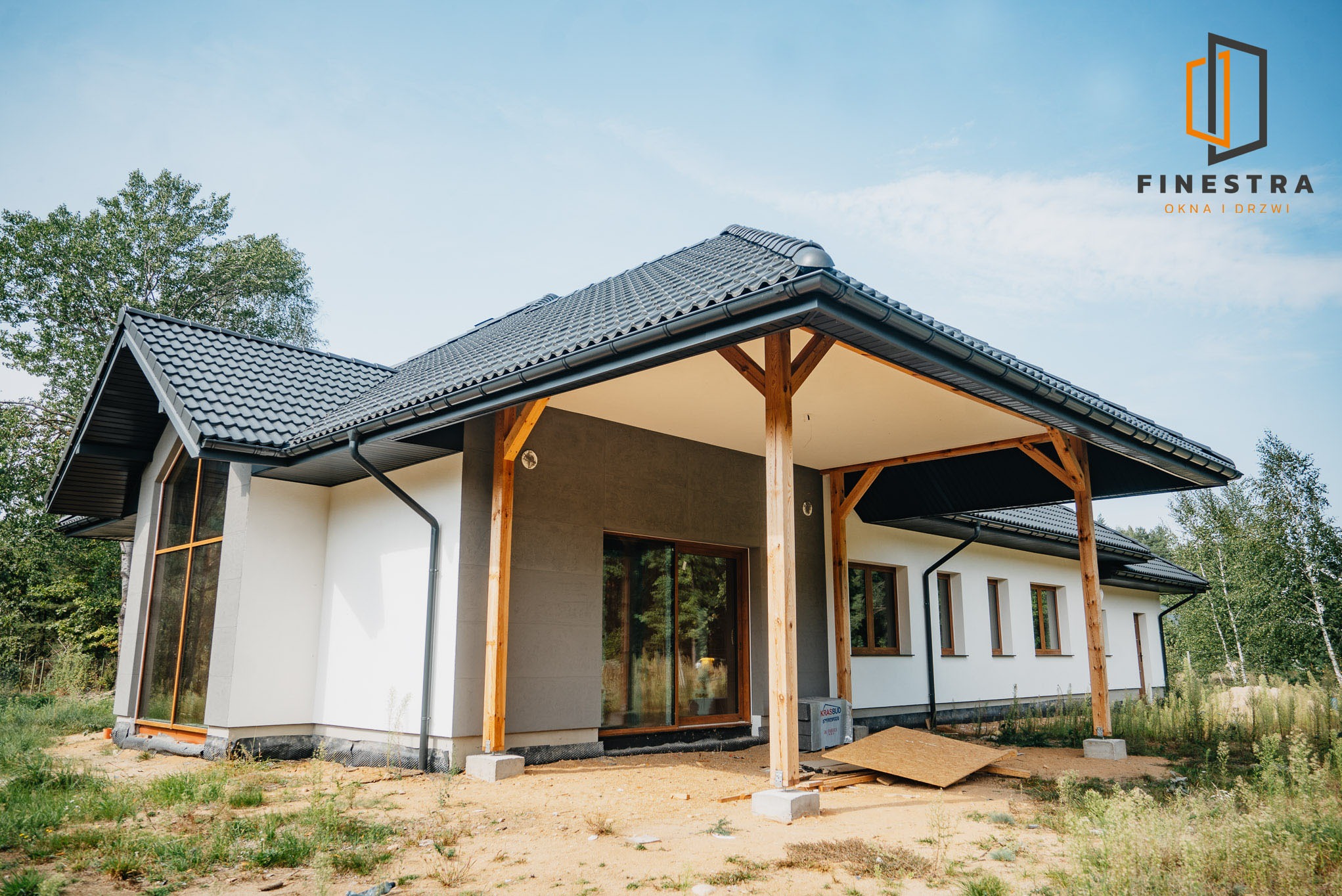 Nowoczesny dom jednorodzinny z dużymi oknami i przesuwnymi drzwiami tarasowymi, wykończony w bieli i szarości, z drewnianą konstrukcją zadaszenia tarasu.